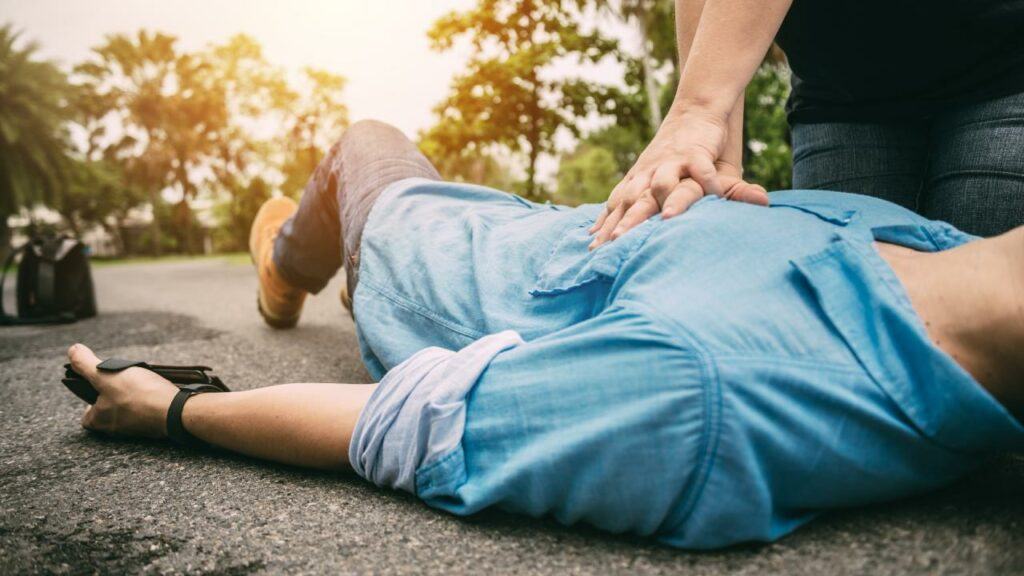 Man in a blue shirt laying on his back with person providing first aid for a heart attack.